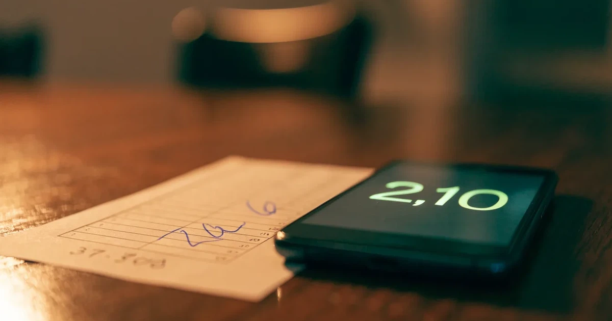 Uma moeda física de bitcoin ao lado de um telemóvel com um talão de aposta no ecrã, sobre uma mesa escura